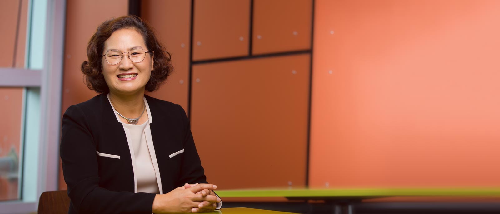 School of Hospitality Business faculty member Mi Ran Kim sits at a table, smiling, against a bright orange backdrop.
