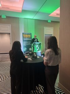 A man stands at a podium delivering remarks at an indoor event, lit by green accent lighting. Two attendees stand in the foreground at a round table, facing the speaker. Behind the podium, a banner reads “Celebrating 100 Years of Hospitality Leadership,” indicating a centennial-themed gathering. The room features neutral walls, exit signs, and a bold geometric patterned carpet.