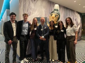 Seven people stand side by side indoors, smiling at the camera in business or business-casual attire. They appear to be at a professional event, with name badges visible on several individuals. Behind them is a decorative wall featuring a large Spartan mascot figure and a sign partially visible, while the floor has a bold geometric patterned carpet.