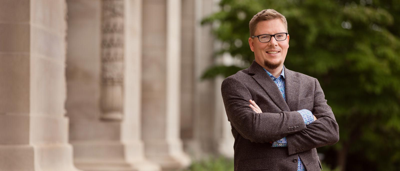 Management faculty member Russ Johnson stands with arms crossed outside on a sunny day, with a stone building on MSU's campus in the background.