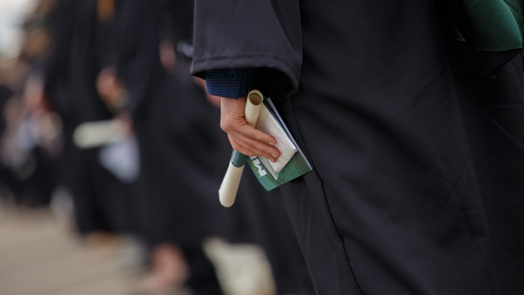 Close up image of an Executive MBA student wearing graduation cap and gown, holding an event program and diploma.