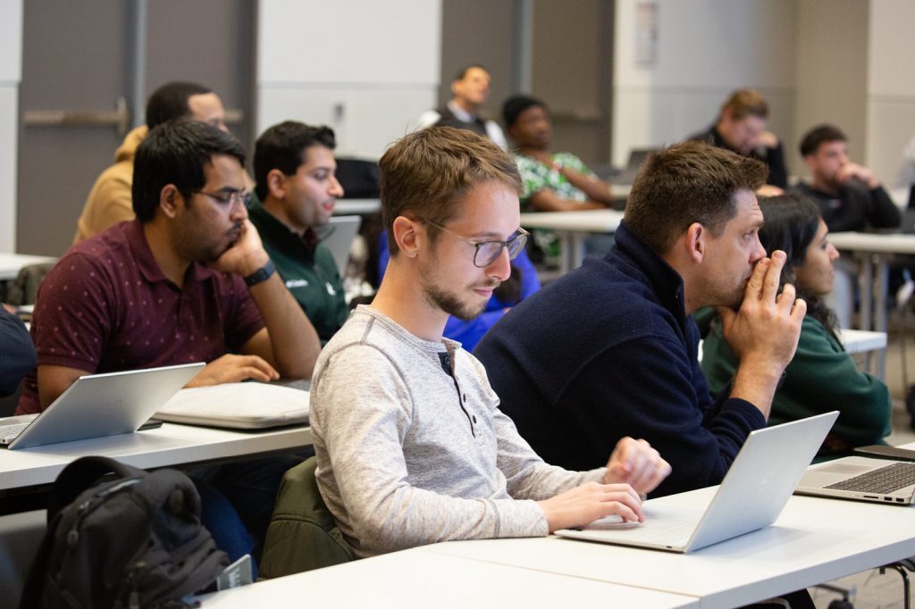 Full-Tima MBA students sit in a classroom, taking notes on laptops while listening to an alumni presentation.