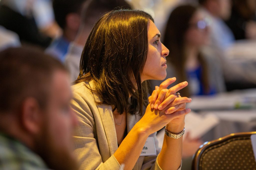 Executive woman in business with hands clasped, gazes to the right in a room of people.