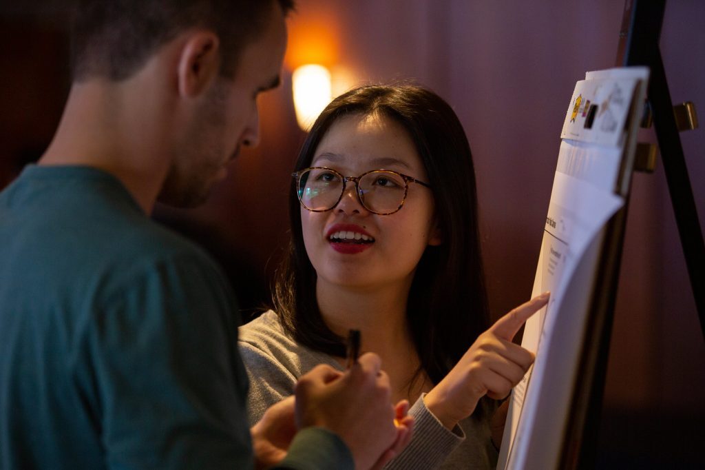 A woman MBA student points at an oversized post-it note while standing and talking to another student.