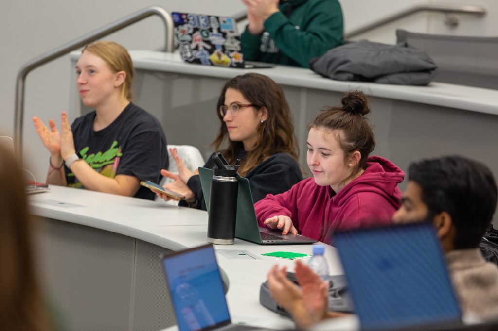 Students clap and take notes on laptops in a classroom during a presentation.