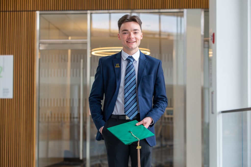 Carlos Conrad stands in the Minskoff Pavilion wearing a suit, holding his graduation cap.