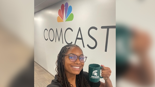 Symone King taking a selfie in front of the Comcast logo holding a Broad College mug