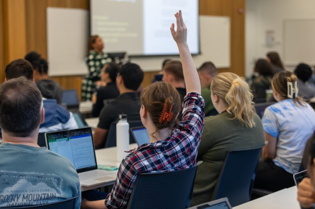 Back row view in a classroom with Dalana Brand speaking. A student raises their hand to ask a question.