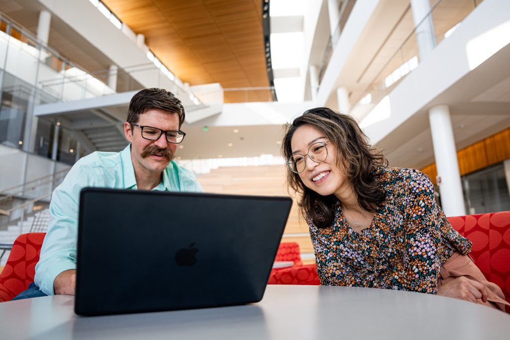 Jason Miller and Li Cheng sit in the Minskoff Pavilion atrium and work on a laptop.