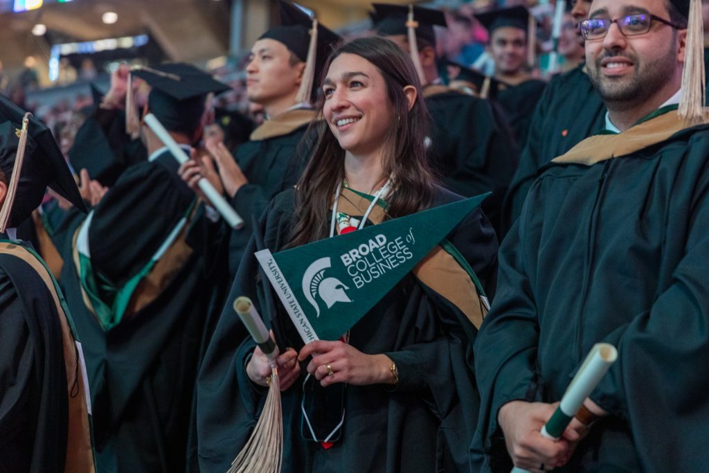 A Spartan wearing advanced degree academic regalia holds a Broad pennant while smiling and standing in the crowd at Breslin Center during MSU Commencement.