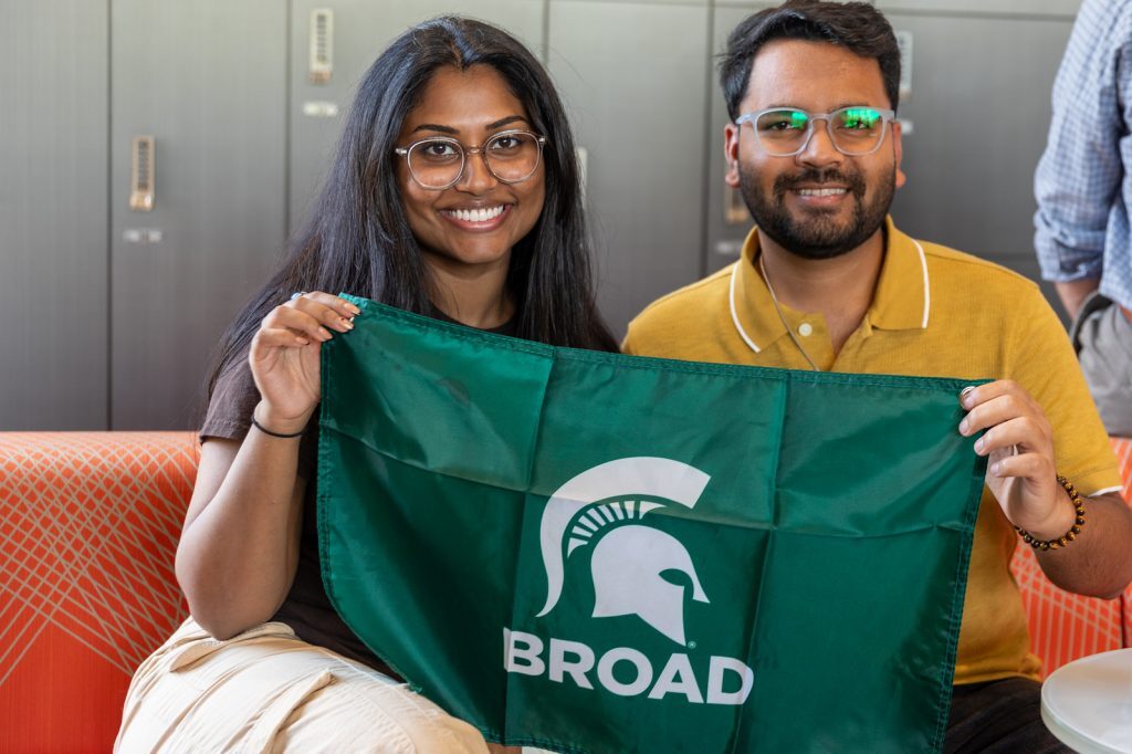 Two Broad Spartans sit and smile while holding a Broad flag.