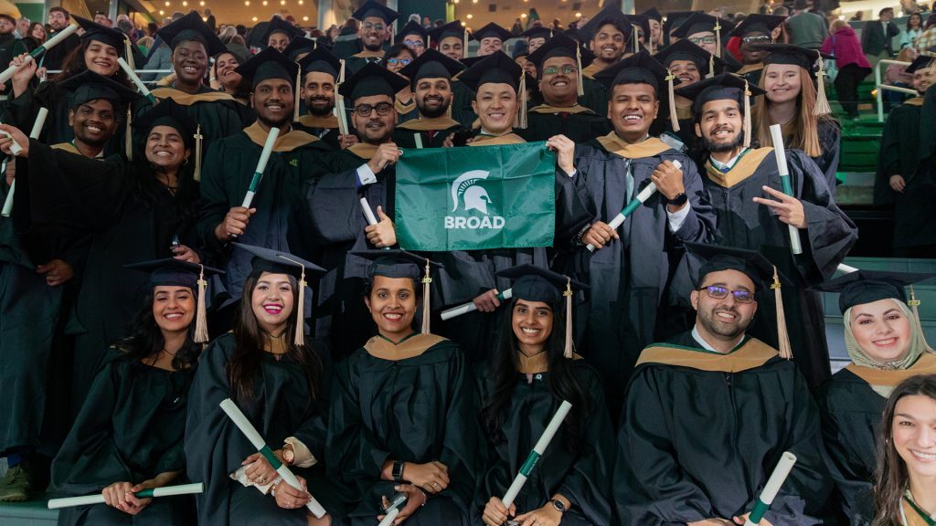 Breslin Center bleachers filled with Broad master's students at commencement, holding diplomas and Broad flags.
