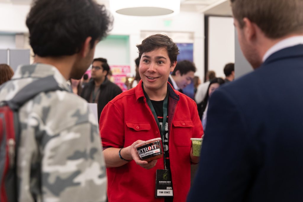 A Spartan entrepreneur holds a Detroit Salsa Company container and smiles while talking with attendees.