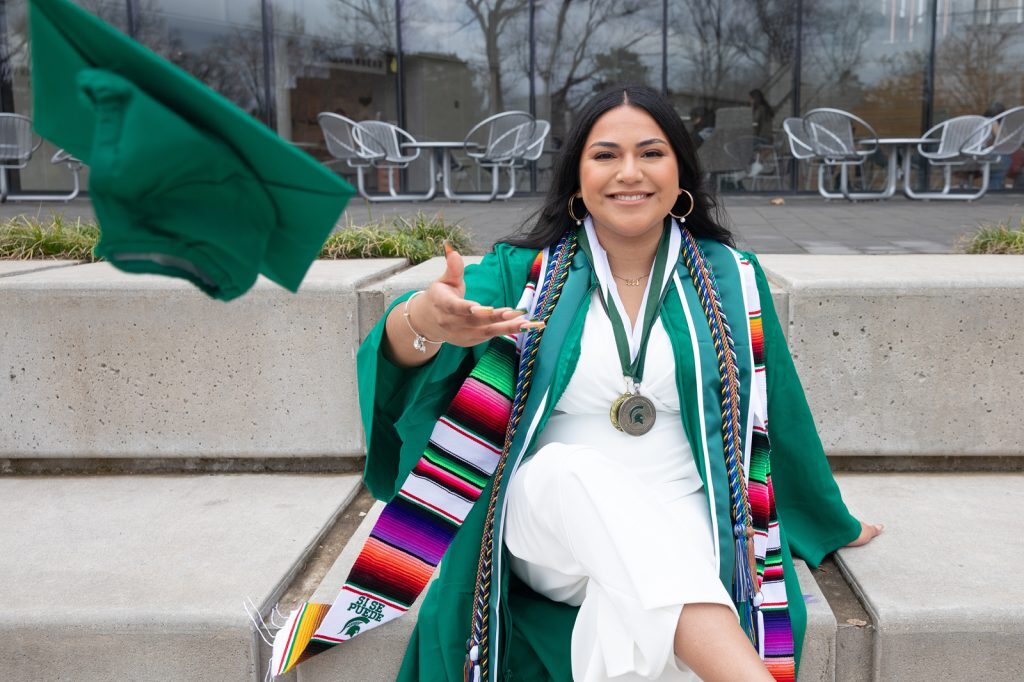 2024 Student commencement speaker Diana Talamantes-Valles wears academic regalia and tosses her cap toward the camera while sitting outside the Minskoff Pavilion.