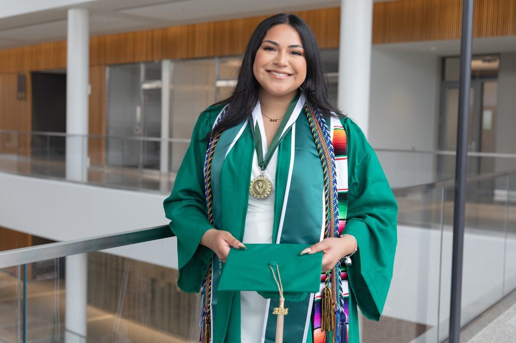 2024 Student commencement speaker Diana Talamantes-Valles wears academic regalia and holds her graduation cap in the Minskoff Pavilion.