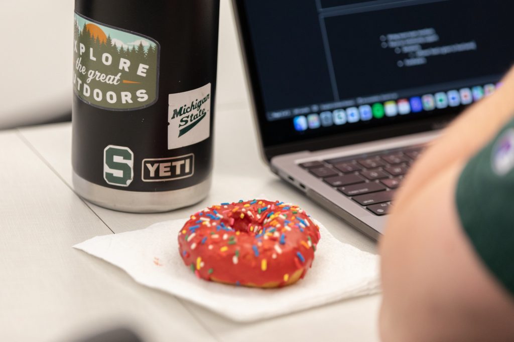 A Groovy Donut with pink icing and sprinkles on napkin near someone with a thermos and laptop.
