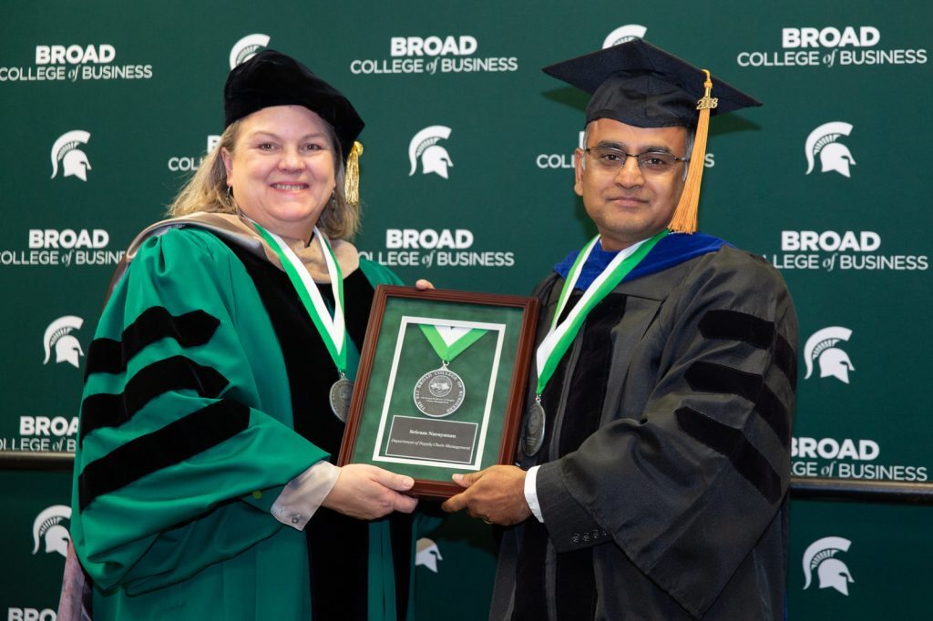 Judith Whipple presents an academic award to Sriram Narayanan at the 2024 Broad College Faculty Investiture Ceremony. They each are wearing academic regalia and stand in front of a Broad-branded backdrop.