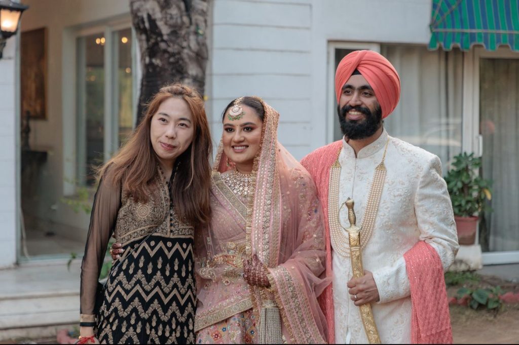 Three people posing at a wedding, including a bride and groom in traditional attire and a woman in a black and gold dress.