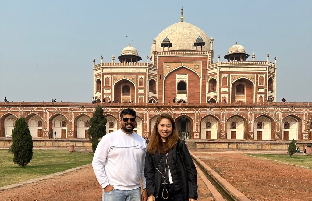 Two people stand on a path in front of a historic building with domes and archways.