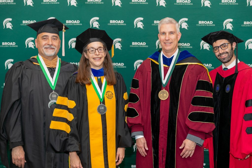 Four people posing in academic regalia in front of a green backdrop that has the Michigan State University Spartan logo and says Broad College of Business.