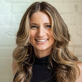Headshot of woman smiling with long wavy hair against a white brick wall.