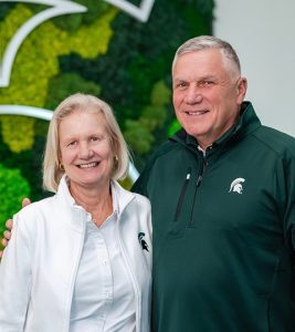 Two people stand side by side in front of a green and black moss wall featuring a large white Michigan State University Spartan helmet design. One person is wearing a white jacket with an MSU logo, and the other is wearing a dark green quarter‑zip pullover with an MSU logo. The person on the right has their arm around the person on the left. The setting appears bright and modern