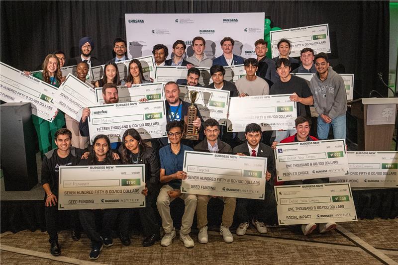 A large group of young adults posing on a stage, smiling and holding oversized ceremonial checks and a trophy, at a startup competition event.