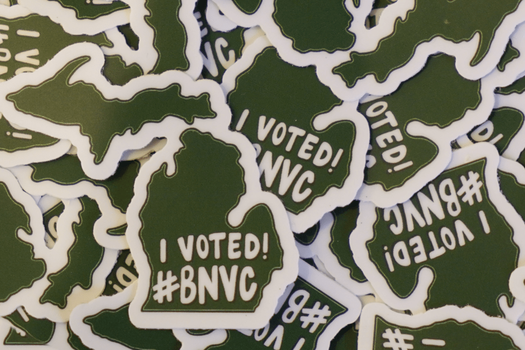 A close-up pile of green stickers shaped like Michigan, each reading “I Voted! #BNVC” in white text.