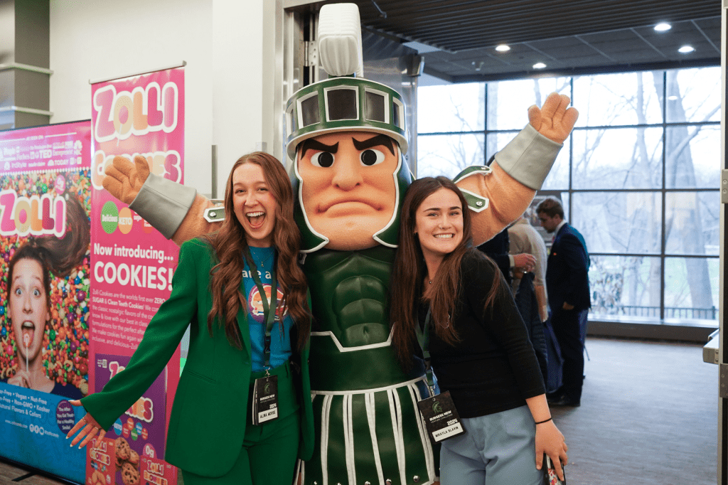 Two smiling women pose with the Michigan State Spartans mascot indoors, arms raised in celebration, with colorful promotional signage and large windows in the background.