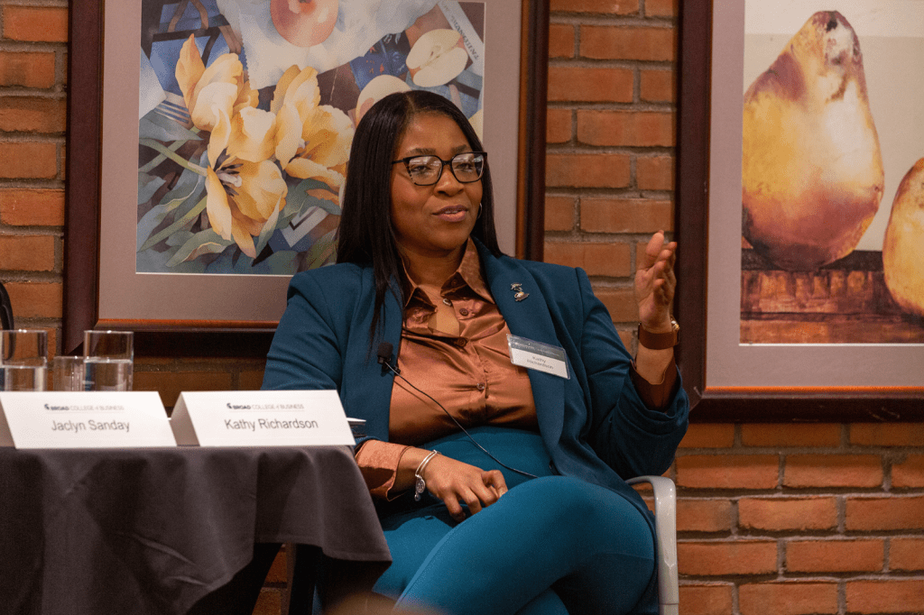 A panelist in a teal suit speaks while seated in front of framed artwork, addressing an audience during a discussion.