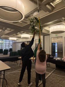 Two people working together to hang a wreath from the ceiling of a ballroom 