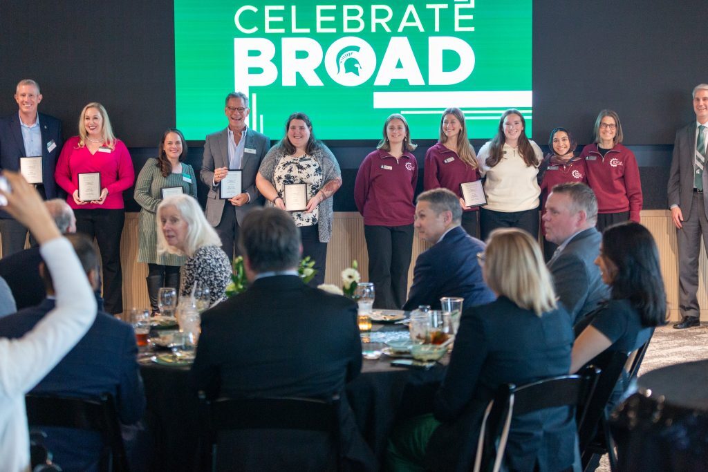 Award recipients stand on stage holding plaques in front of a large “Celebrate Broad” screen as attendees watch from banquet tables.