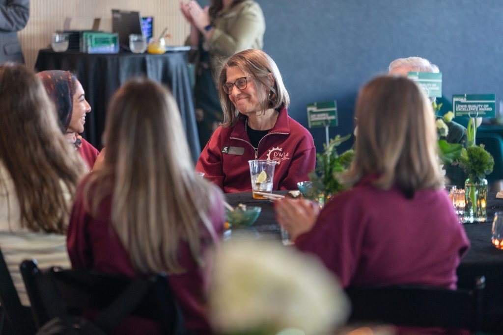 A woman seated at a banquet table smiles and applauds, surrounded by fellow attendees wearing matching maroon quarter-zips.