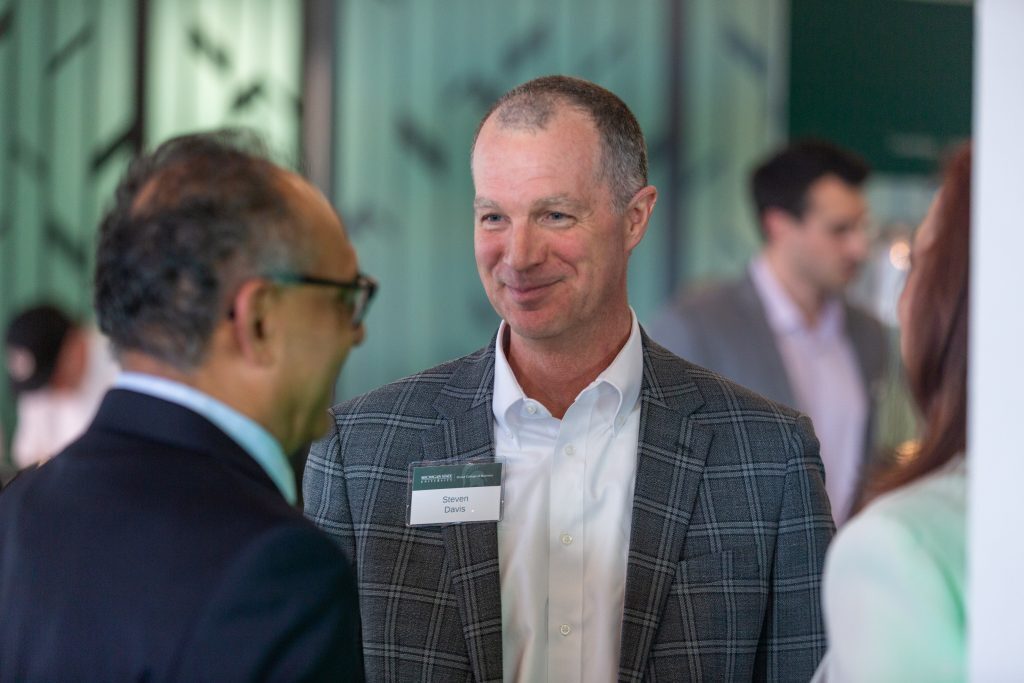 Two attendees smile and chat during a networking reception, standing among other guests in a bright event space.