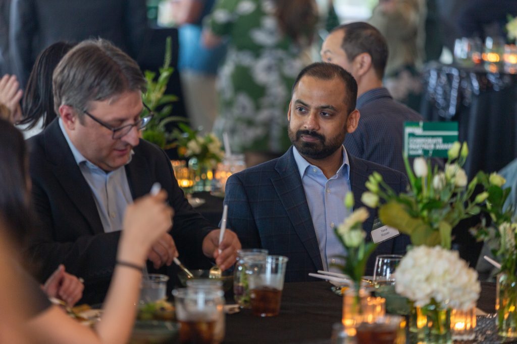 Two attendees seated at a banquet table converse during the event, with floral centerpieces and candlelight visible in the foreground.