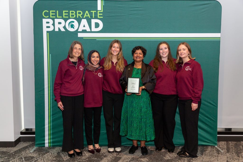 An award recipient holding a plaque poses with a group of students in matching maroon quarter-zips in front of a “Celebrate Broad” backdrop.