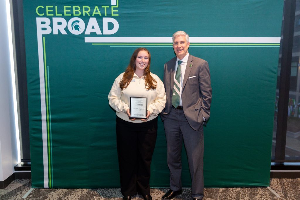 A student holding an award plaque poses with a college leader in front of a green “Celebrate Broad” backdrop.