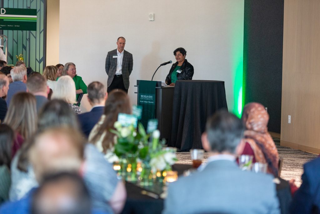 A woman speaks at a podium during the event while attendees seated at round tables listen.