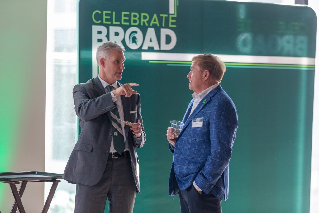 Two men in suits converse in front of a large “Celebrate Broad” display during a reception.