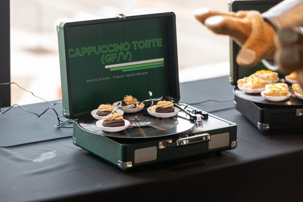 Small plated desserts are displayed on a record player-themed serving station with a sign reading “Cappuccino Torte.”
