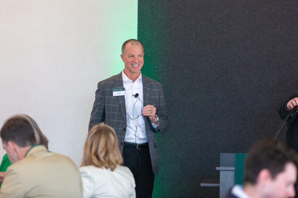 A man in a plaid blazer smiles while speaking to attendees during the event.