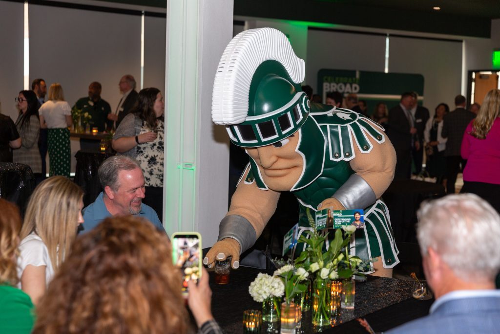 The Michigan State Spartan mascot leans toward a table of guests, interacting playfully during the celebration reception.