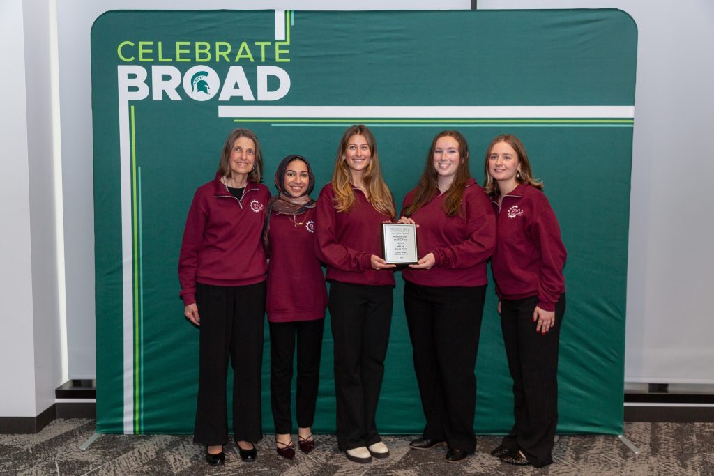 An award recipient holding a plaque poses with a group of students in matching maroon quarter-zips in front of a “Celebrate Broad” backdrop.