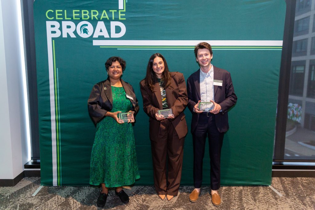 Three alumni stand in front of a green “Celebrate Broad” backdrop, each holding a glass award and smiling toward the camera.