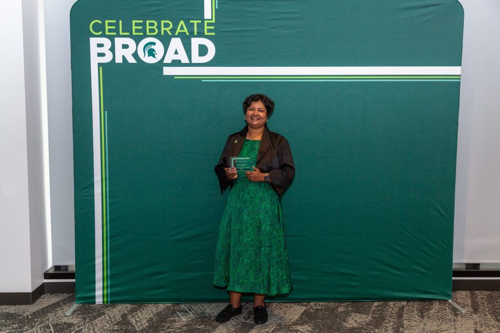 An award recipient in a green dress and black jacket stands in front of a “Celebrate Broad” backdrop, holding a glass award.