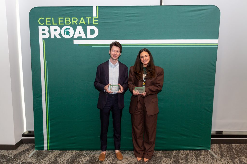 Two alumni stand side by side in front of a “Celebrate Broad” backdrop, each holding a glass award.