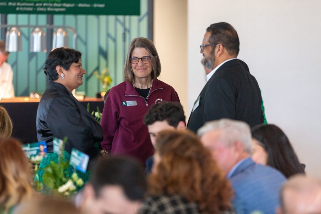 Three attendees stand together in conversation during the Celebrate Broad event while guests mingle in the background.