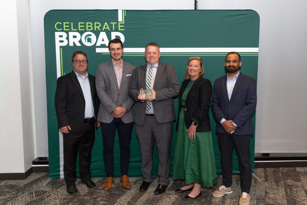 Five award recipients stand together in front of a “Celebrate Broad” backdrop, with the center recipient holding a glass award.