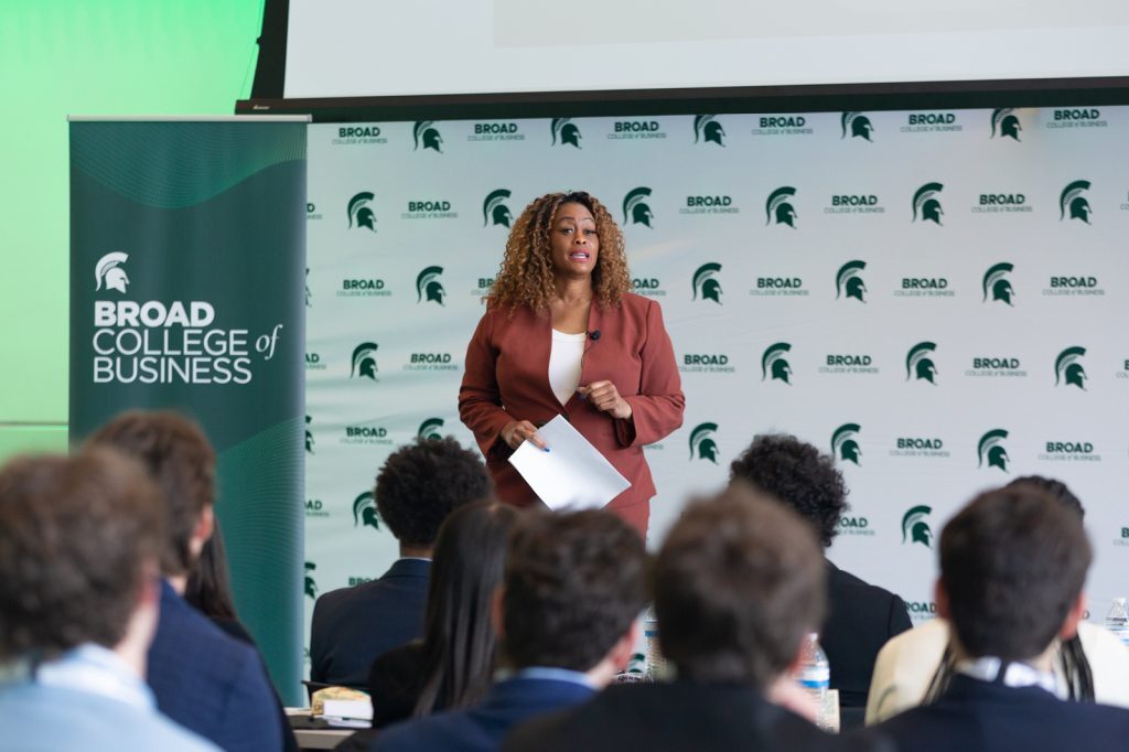 Speaker addresses an audience at the Broad College of Business, standing in front of a branded backdrop and holding notes.