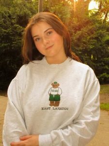 Person standing outdoors at sunset wearing a light gray sweatshirt with an “East Lansing” graphic, smiling softly with trees and warm sunlight in the background.